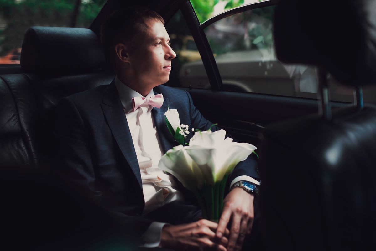 handsome groom sits with wedding bouquet inside a car and look in window outside handsome groom sits with wedding bouquet inside a car and look in window outside